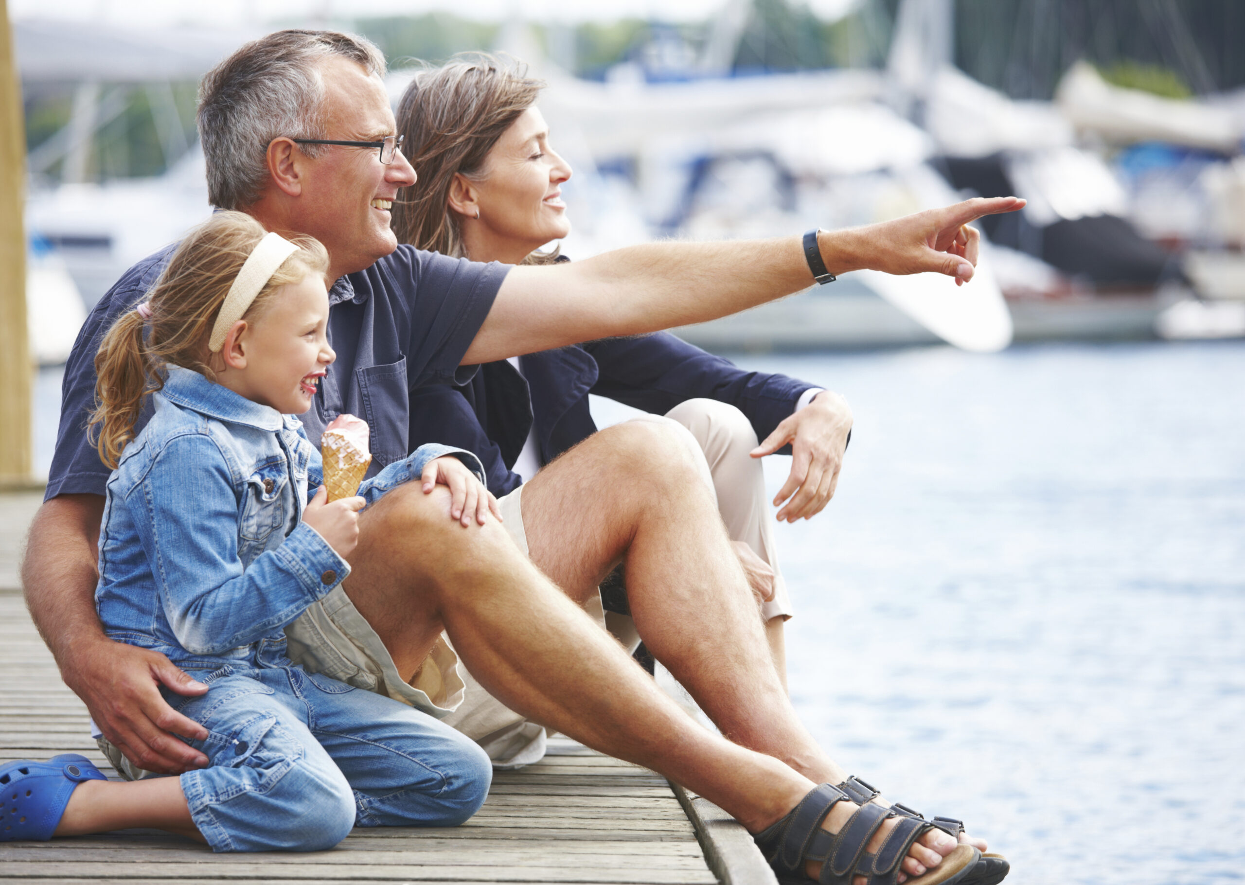 Happy Grandparents sitting with their granddaughter on a pier, pointing out at the boats - copyspace